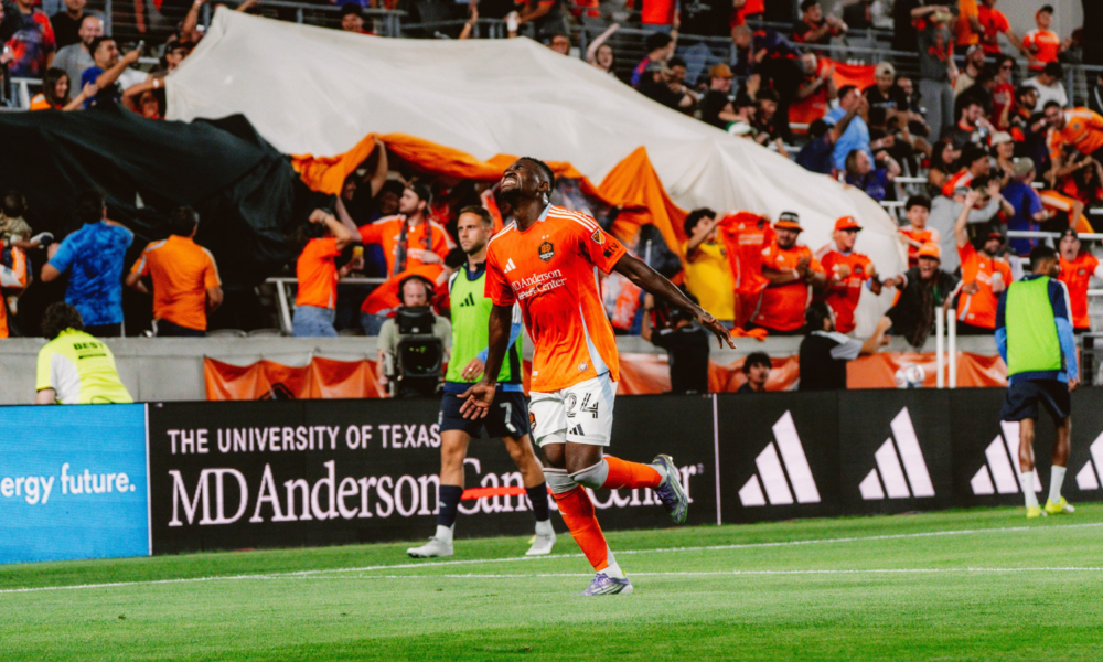 Soccer player in orange uniform celebrates a goal on the field with cheering fans in the stands behind him