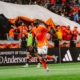 Soccer player in orange uniform celebrates a goal on the field with cheering fans in the stands behind him