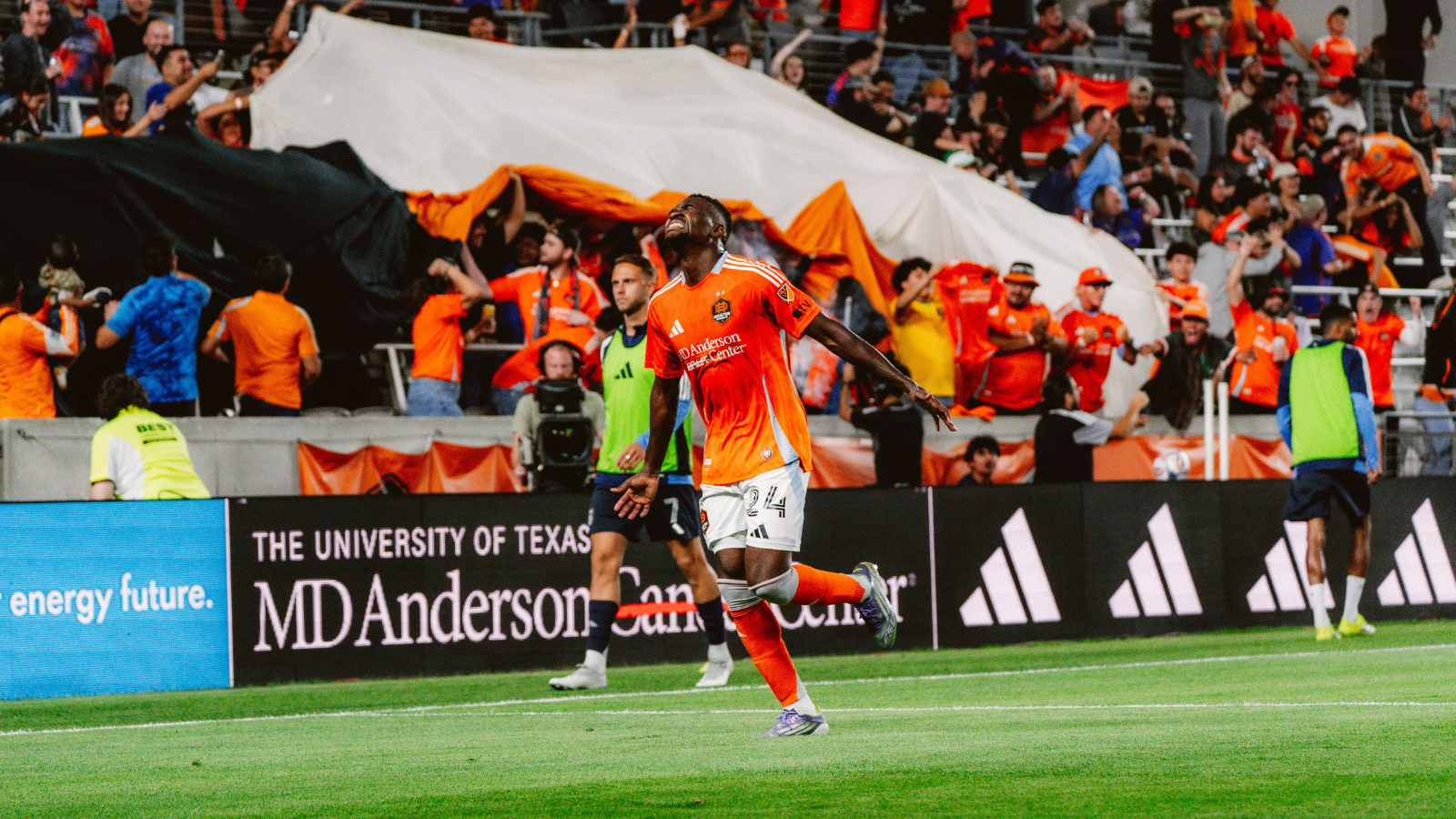 Soccer player in orange uniform celebrates a goal on the field with cheering fans in the stands behind him