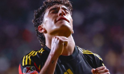 Soccer player in a black jersey with yellow stripes celebrating with clenched fists in a stadium crowd behind him