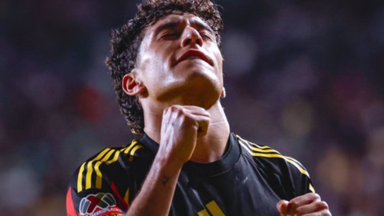 Soccer player in a black jersey with yellow stripes celebrating with clenched fists in a stadium crowd behind him