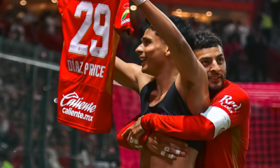 Two celebrating players embrace on the field, one lifting a red jersey with number 29 and sponsor branding visible, crowd in the background.