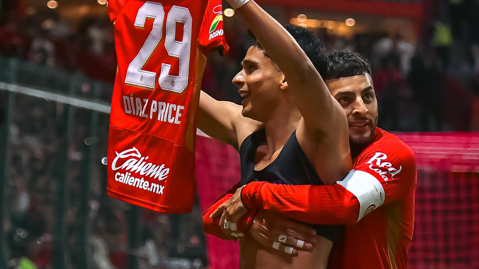 Two celebrating players embrace on the field, one lifting a red jersey with number 29 and sponsor branding visible, crowd in the background.