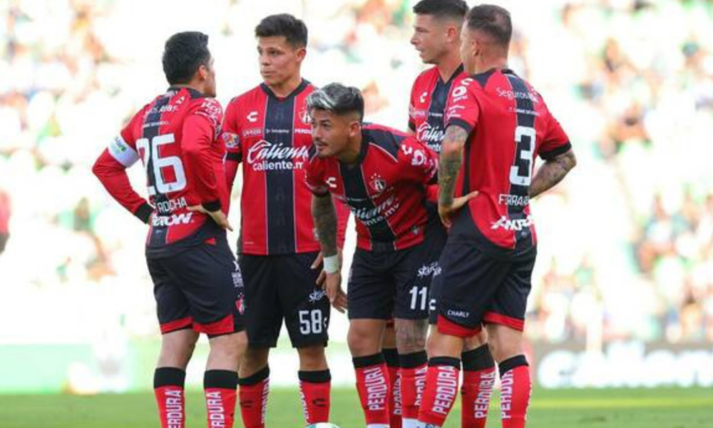 Five soccer players in red-and-black striped uniforms huddle on a green pitch during a match.