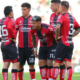 Five soccer players in red-and-black striped uniforms huddle on a green pitch during a match.