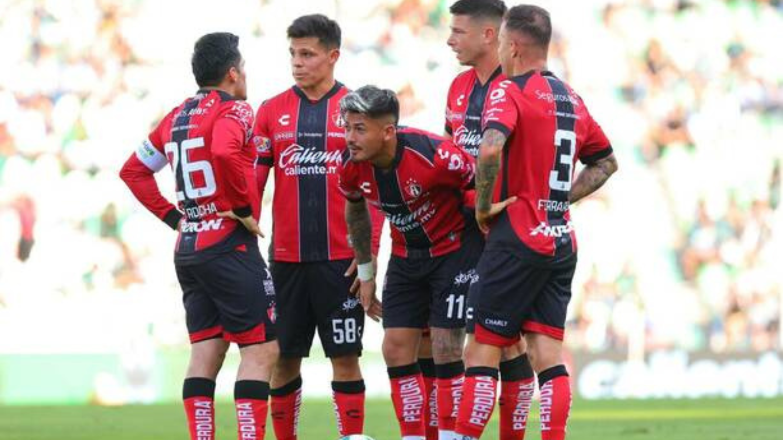 Five soccer players in red-and-black striped uniforms huddle on a green pitch during a match.