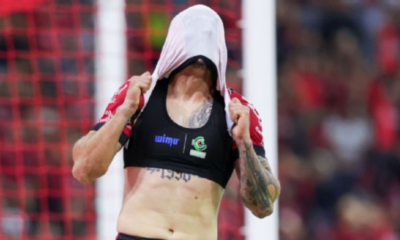 Female athlete pulls a shirt over her head to reveal a black sports bra with logos, tattoos visible on her arm, on a soccer field with a goal net in the background