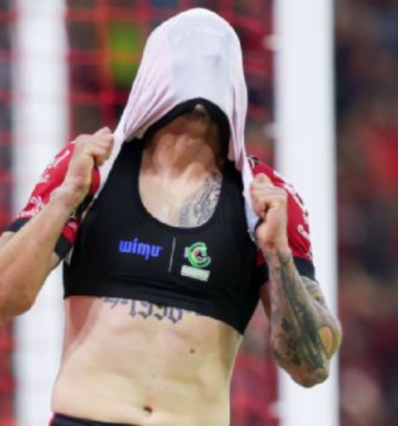 Female athlete pulls a shirt over her head to reveal a black sports bra with logos, tattoos visible on her arm, on a soccer field with a goal net in the background