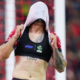 Female athlete pulls a shirt over her head to reveal a black sports bra with logos, tattoos visible on her arm, on a soccer field with a goal net in the background