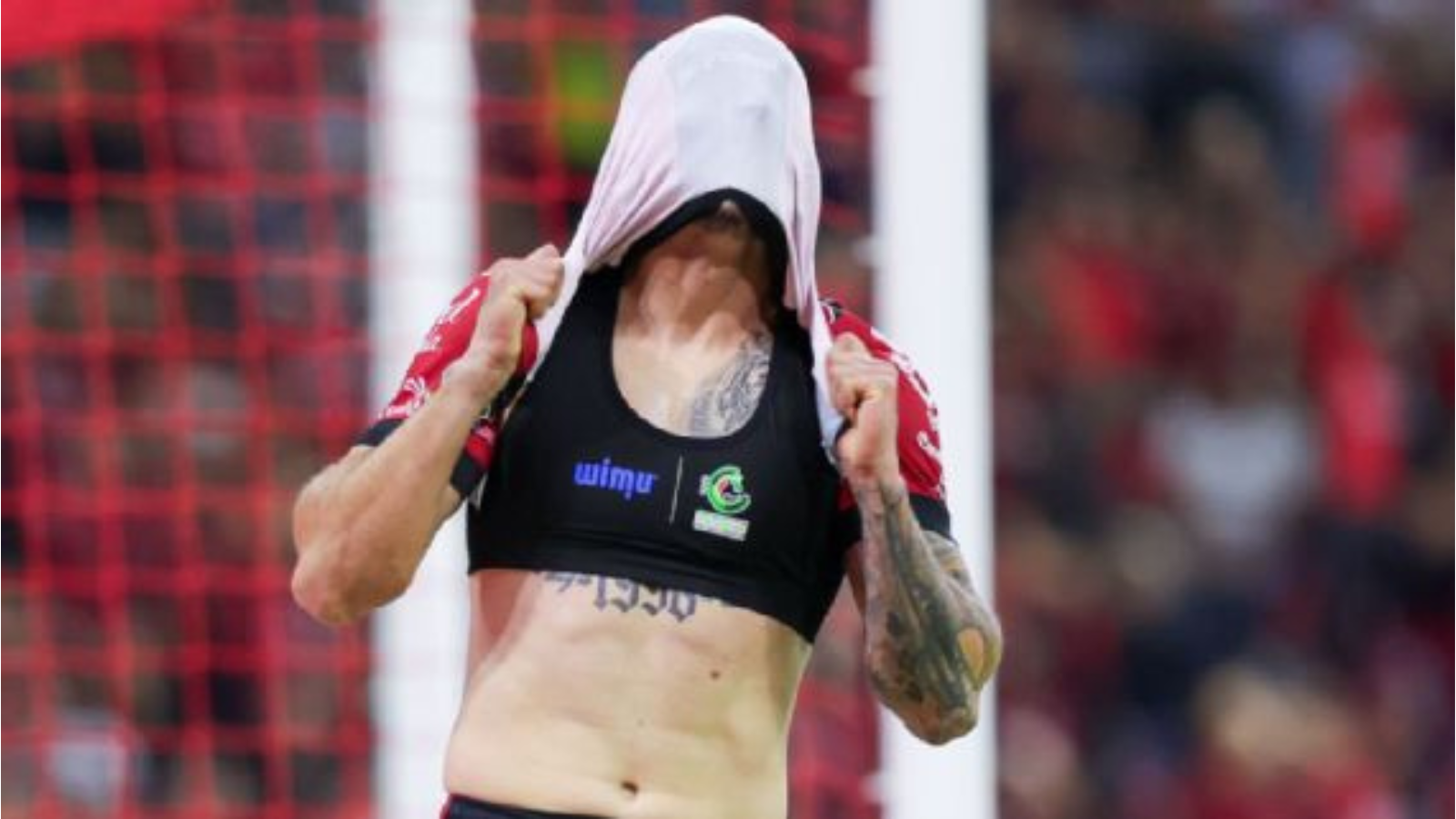Female athlete pulls a shirt over her head to reveal a black sports bra with logos, tattoos visible on her arm, on a soccer field with a goal net in the background