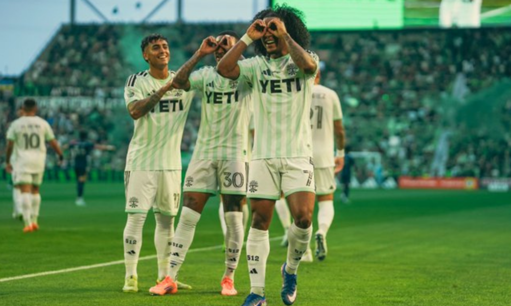 Soccer players in light green uniforms celebrate on the field, making heart shapes with hands amid a cheering crowd.
