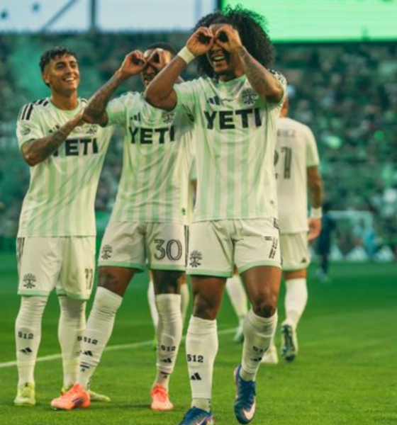 Soccer players in light green uniforms celebrate on the field, making heart shapes with hands amid a cheering crowd.