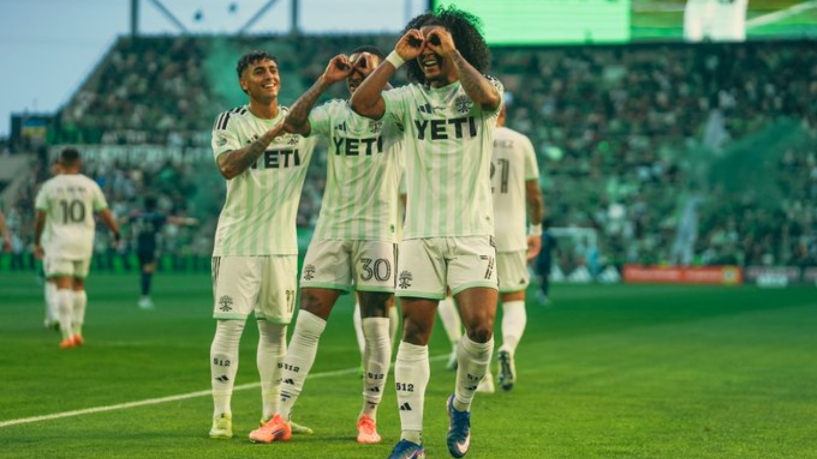 Soccer players in light green uniforms celebrate on the field, making heart shapes with hands amid a cheering crowd.