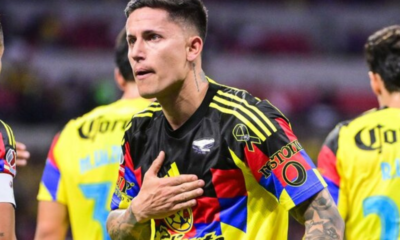 Soccer player in a colorful jersey places hand over heart during a match, with teammates in yellow uniforms in the background.