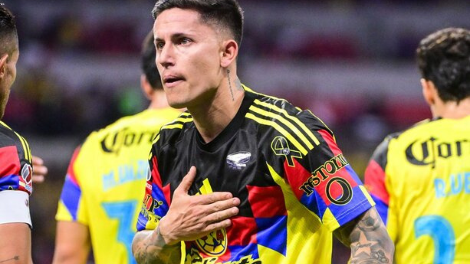 Soccer player in a colorful jersey places hand over heart during a match, with teammates in yellow uniforms in the background.