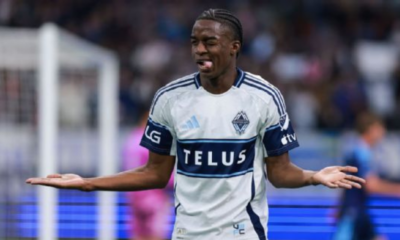 Soccer player in white and blue jersey with TELUS sponsor, arms outstretched in confusion on the field.