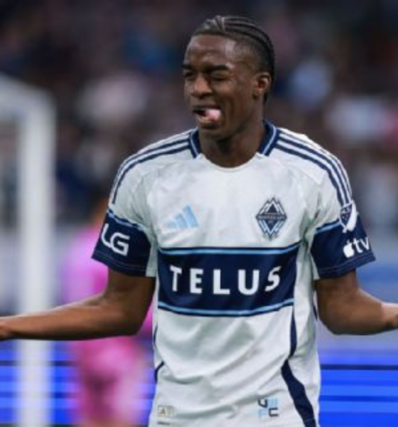 Soccer player in white and blue jersey with TELUS sponsor, arms outstretched in confusion on the field.