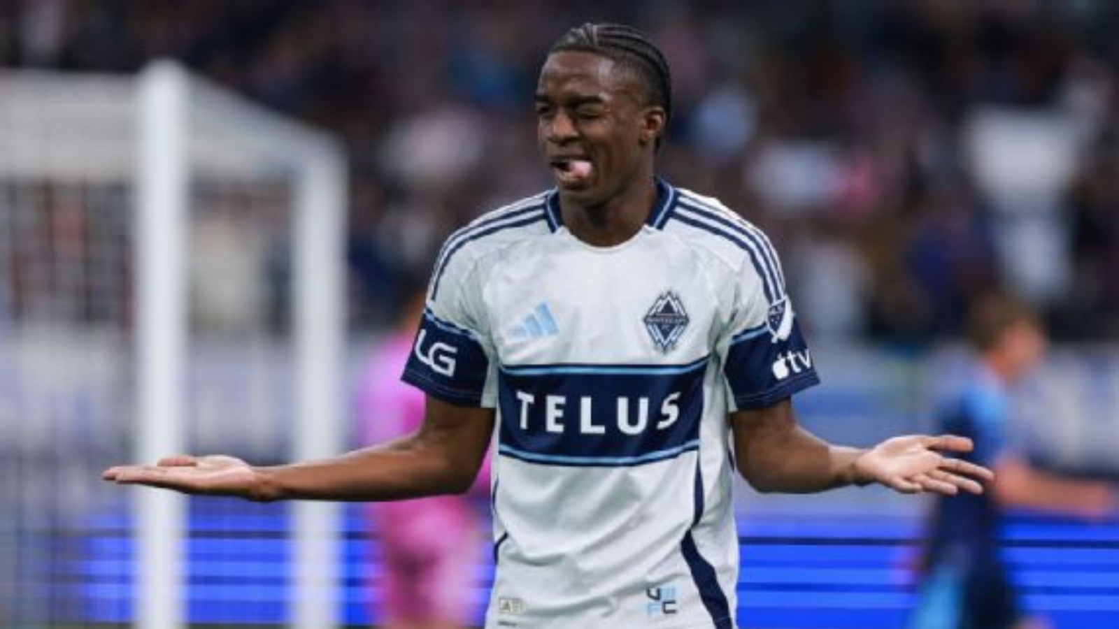 Soccer player in white and blue jersey with TELUS sponsor, arms outstretched in confusion on the field.