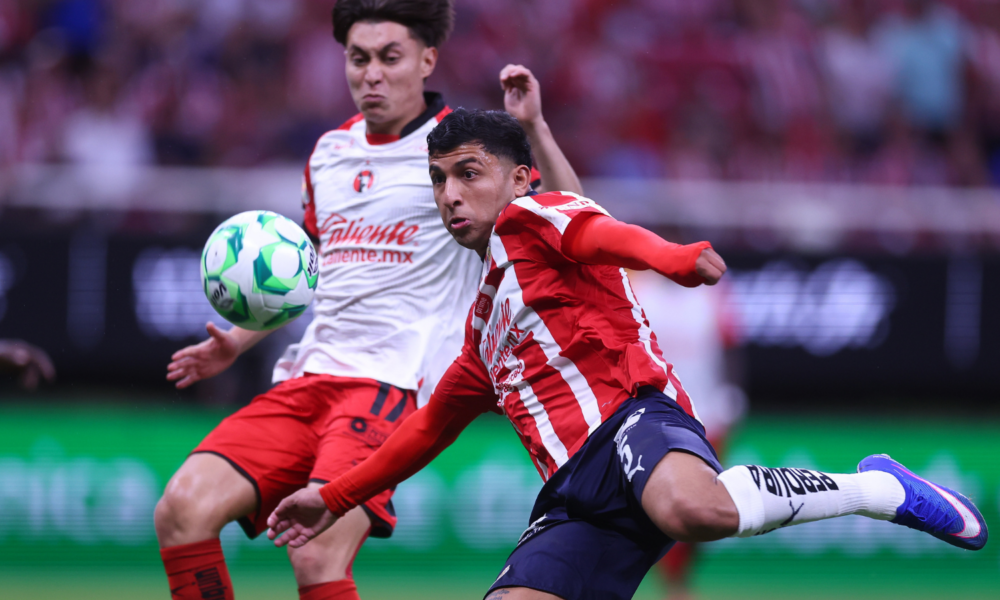 Two soccer players contest a ball mid-action on the field, one in a red striped jersey and the other in a white kit with red shorts, as spectators watch in the background.