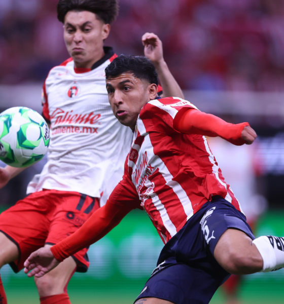 Two soccer players contest a ball mid-action on the field, one in a red striped jersey and the other in a white kit with red shorts, as spectators watch in the background.