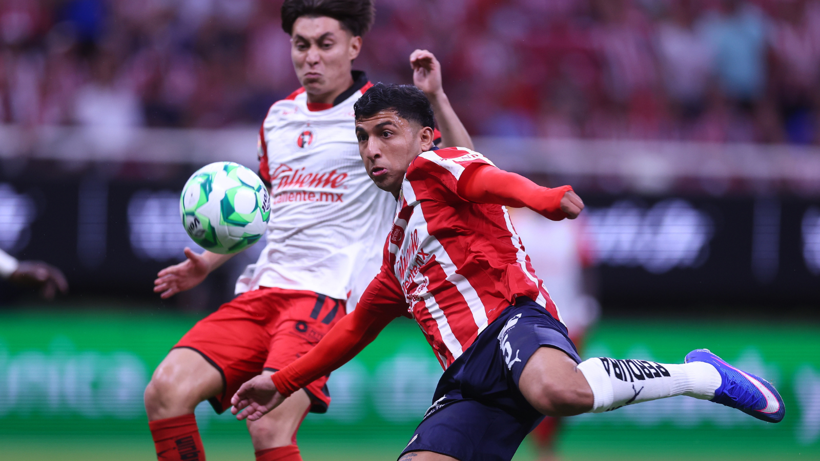 Two soccer players contest a ball mid-action on the field, one in a red striped jersey and the other in a white kit with red shorts, as spectators watch in the background.