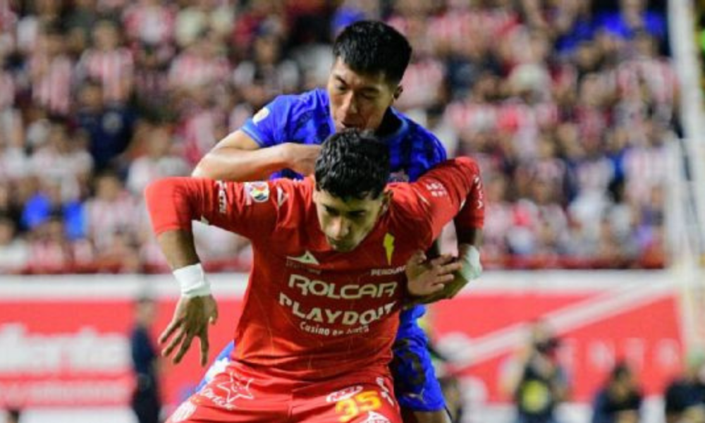 Two soccer players clash for the ball in a crowded stadium, one in red jersey in the foreground and a blue-jersey opponent behind him.