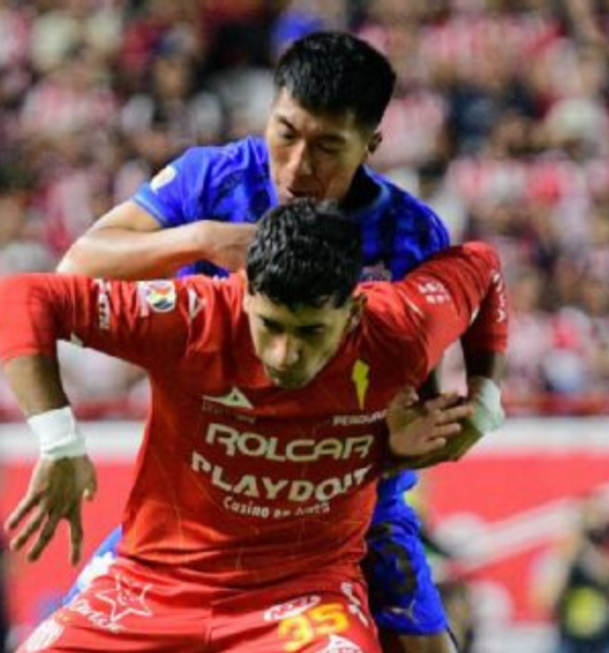 Two soccer players clash for the ball in a crowded stadium, one in red jersey in the foreground and a blue-jersey opponent behind him.