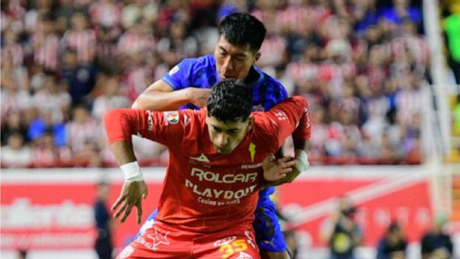 Two soccer players clash for the ball in a crowded stadium, one in red jersey in the foreground and a blue-jersey opponent behind him.