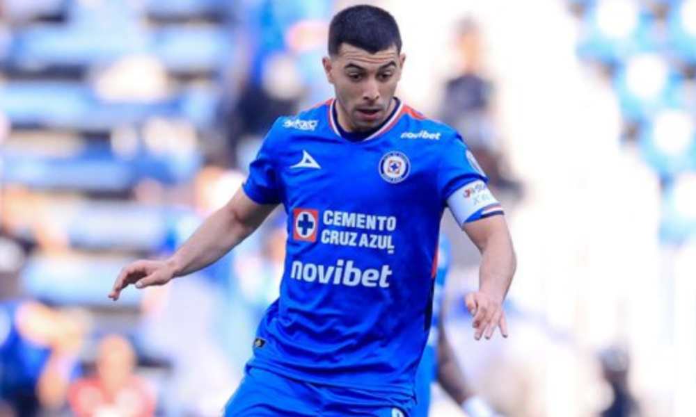 Soccer player in blue kit running with the ball during a match, captain's armband visible.
