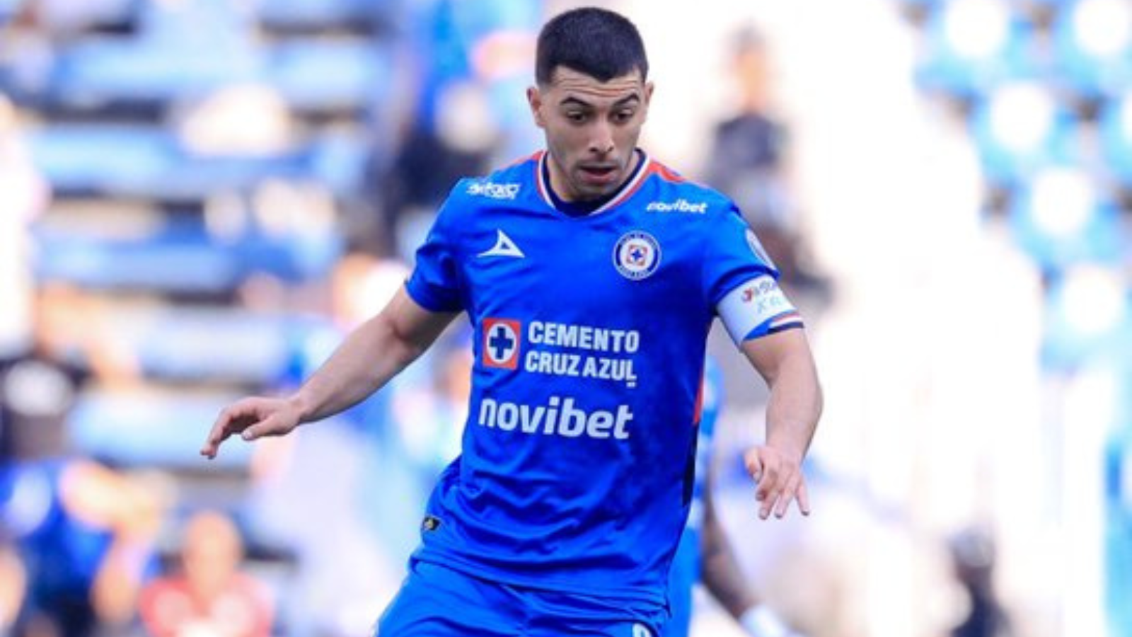 Soccer player in blue kit running with the ball during a match, captain's armband visible.
