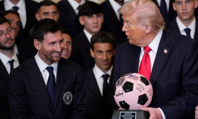 Smiling man in a dark suit and blue tie sits beside Donald Trump, who holds a pink and black signed soccer ball, while a group of suited men watches.