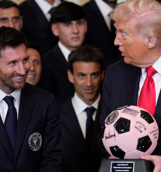 Smiling man in a dark suit and blue tie sits beside Donald Trump, who holds a pink and black signed soccer ball, while a group of suited men watches.