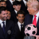 Smiling man in a dark suit and blue tie sits beside Donald Trump, who holds a pink and black signed soccer ball, while a group of suited men watches.