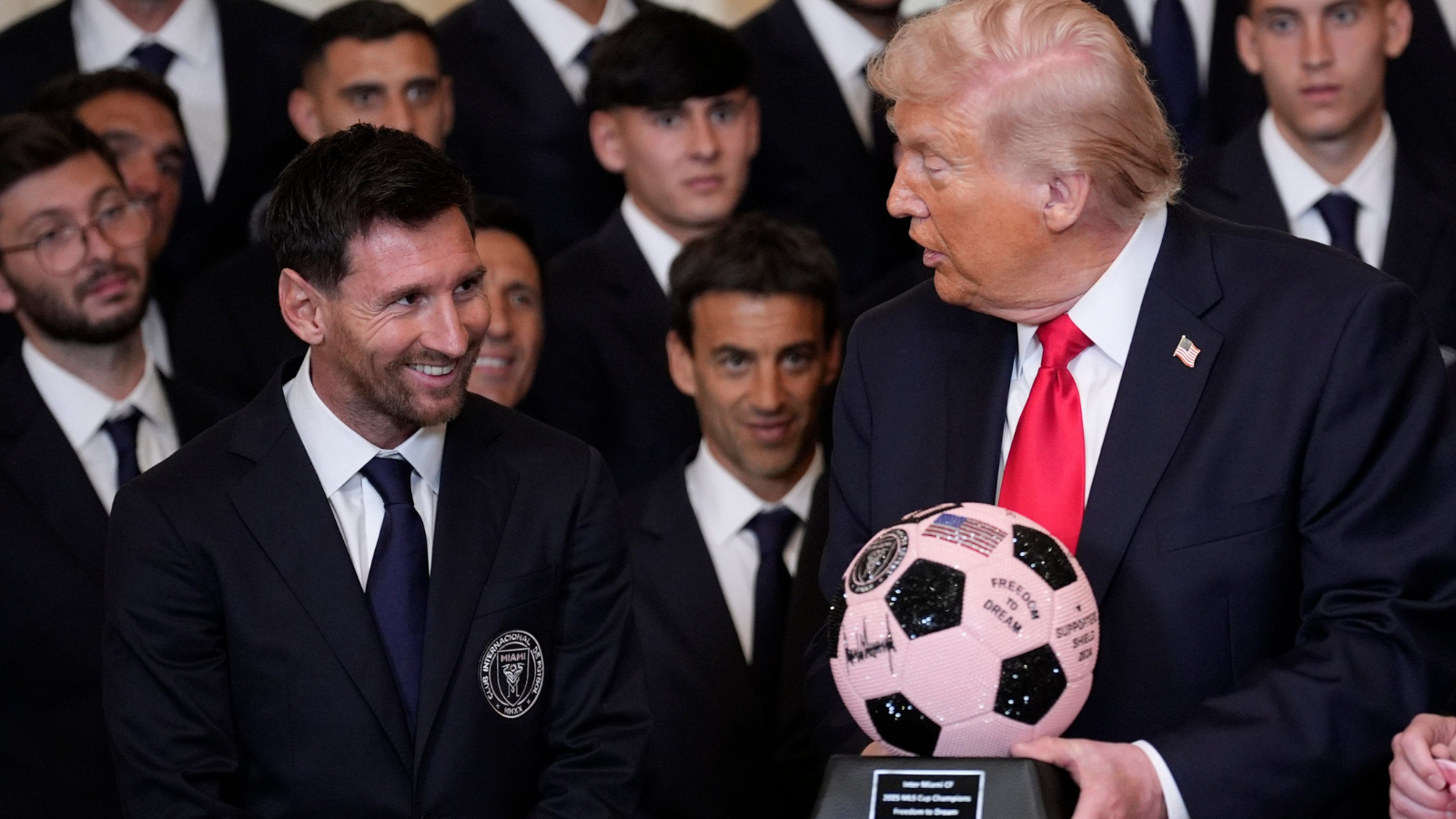 Smiling man in a dark suit and blue tie sits beside Donald Trump, who holds a pink and black signed soccer ball, while a group of suited men watches.