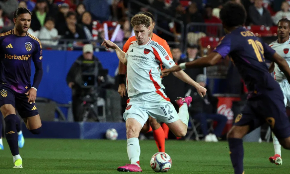 Soccer player in a white kit kicking a ball during a match, surrounded by opposing players in dark purple uniforms.