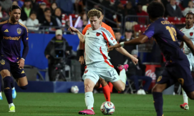 Soccer player in a white kit kicking a ball during a match, surrounded by opposing players in dark purple uniforms.