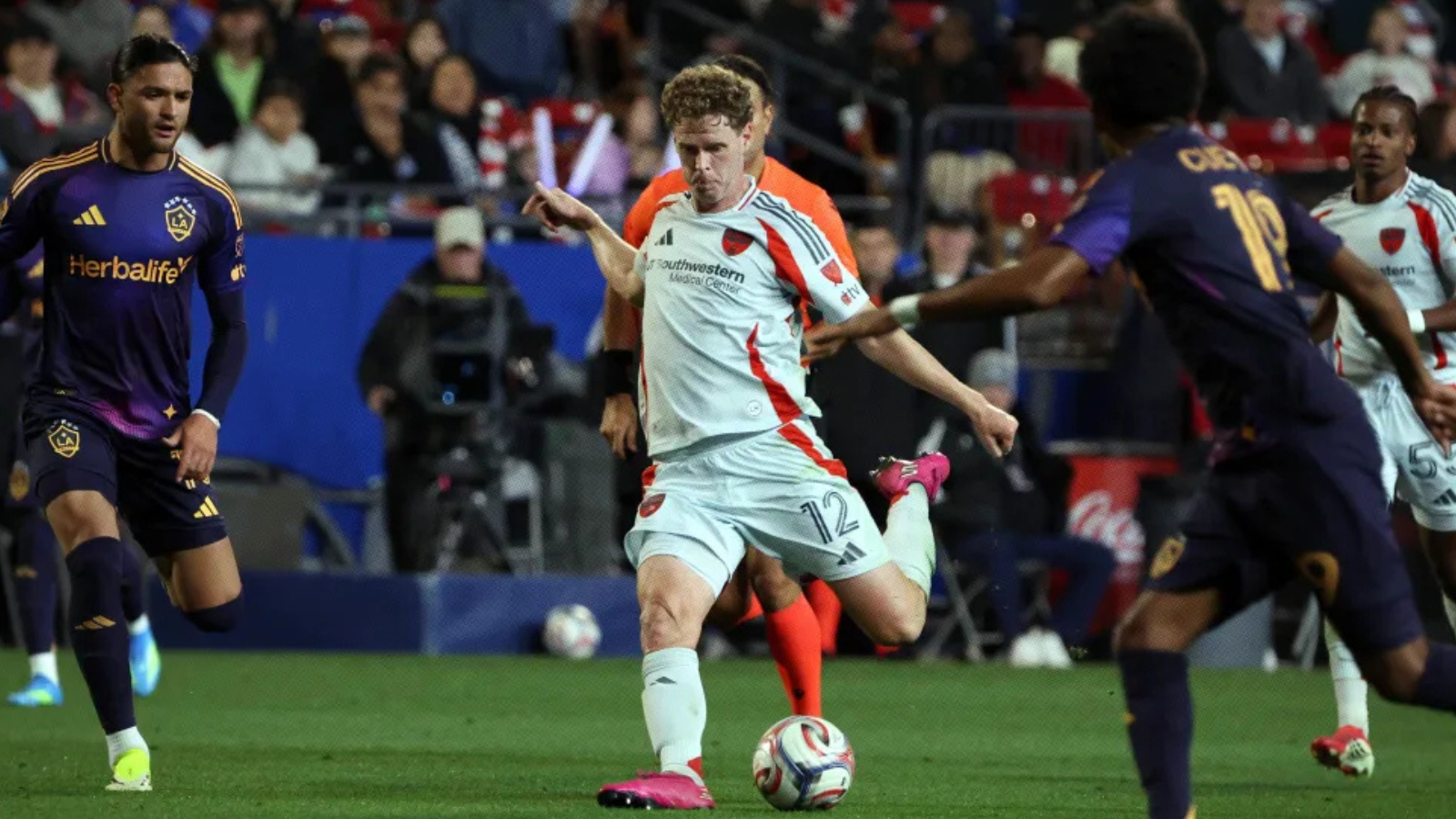 Soccer player in a white kit kicking a ball during a match, surrounded by opposing players in dark purple uniforms.