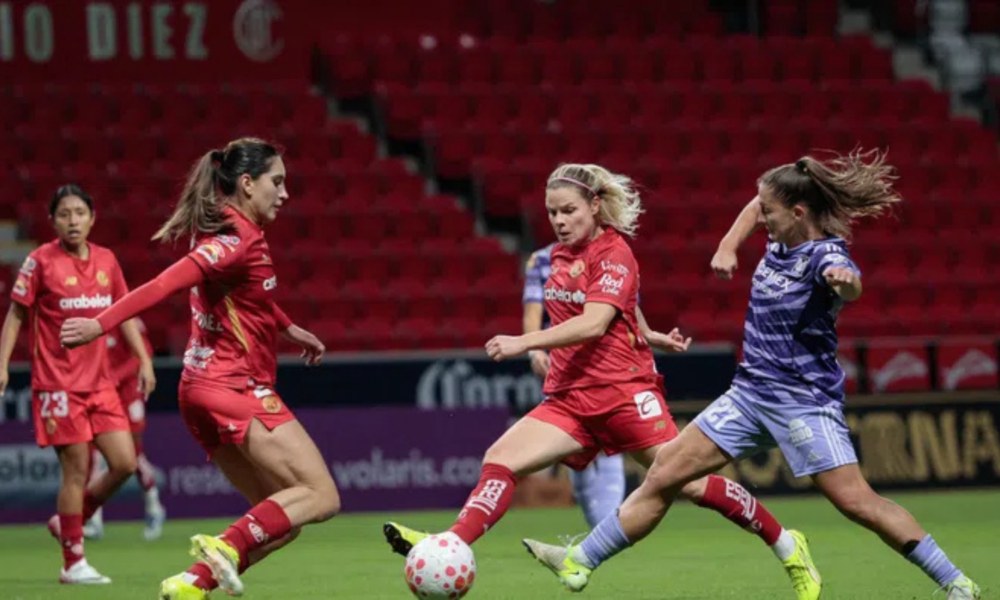 Four female soccer players battle for the ball on a green pitch; three wear red uniforms and one wears purple.