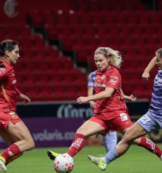 Four female soccer players battle for the ball on a green pitch; three wear red uniforms and one wears purple.