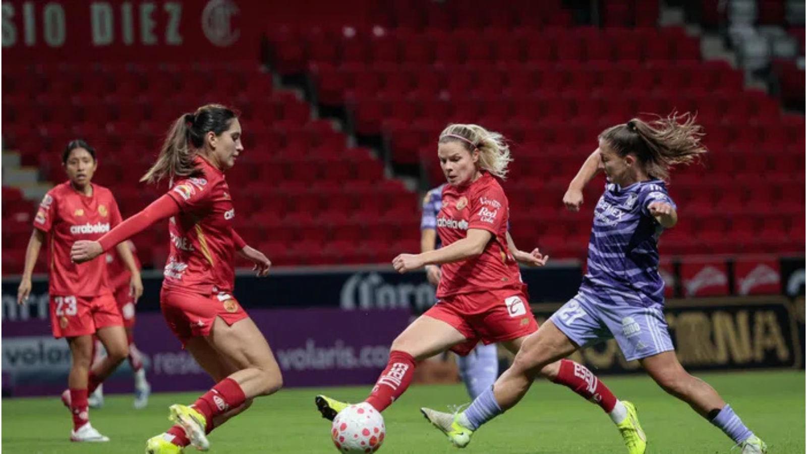 Four female soccer players battle for the ball on a green pitch; three wear red uniforms and one wears purple.