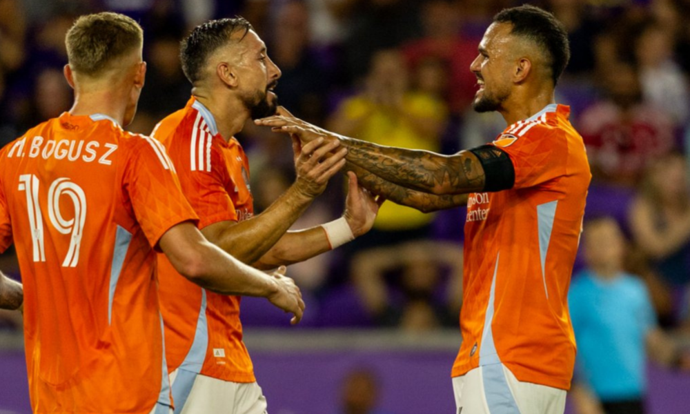 Soccer players in orange jerseys celebrate with a joint high-five on the pitch during a match.