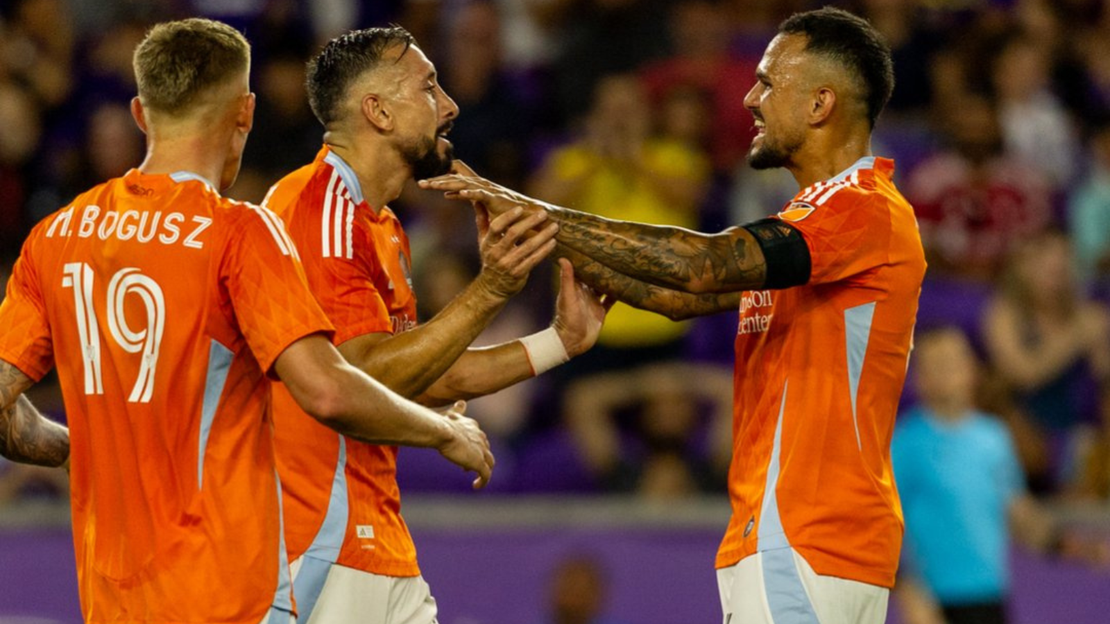 Soccer players in orange jerseys celebrate with a joint high-five on the pitch during a match.