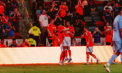 Two players in red jerseys hug on the field as teammates celebrate nearby with a cheering crowd in the stands behind them.