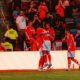 Two players in red jerseys hug on the field as teammates celebrate nearby with a cheering crowd in the stands behind them.