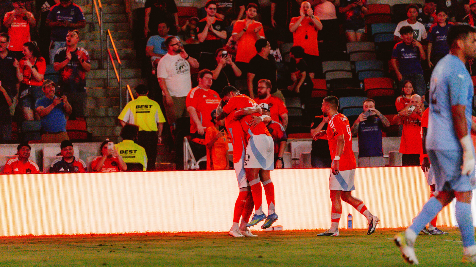 Two players in red jerseys hug on the field as teammates celebrate nearby with a cheering crowd in the stands behind them.