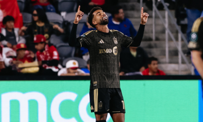 Soccer player in a black and gold LAFC kit raises both arms in celebration on the pitch, fans in the stands behind him.