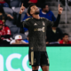 Soccer player in a black and gold LAFC kit raises both arms in celebration on the pitch, fans in the stands behind him.