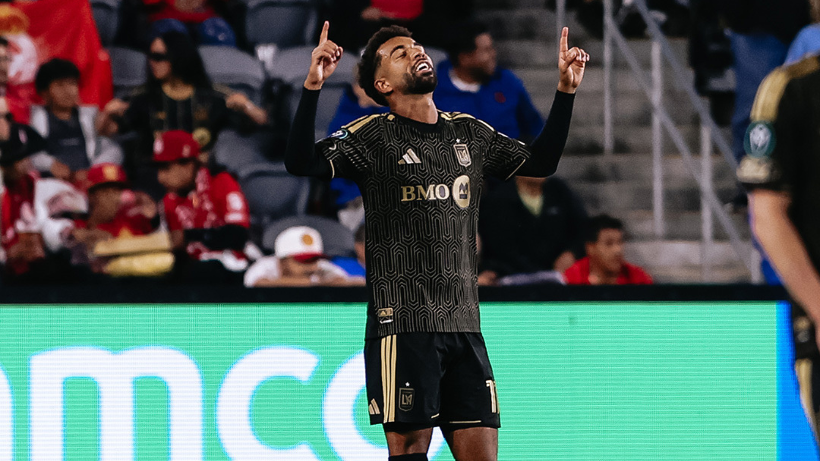 Soccer player in a black and gold LAFC kit raises both arms in celebration on the pitch, fans in the stands behind him.