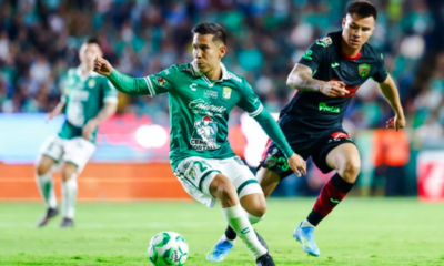 Two soccer players chase a green-and-white ball on the field during a match, with a blurred crowd in the background.