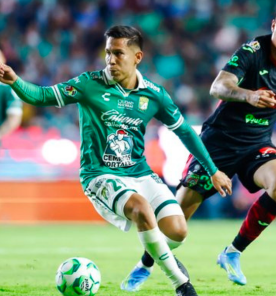 Two soccer players chase a green-and-white ball on the field during a match, with a blurred crowd in the background.